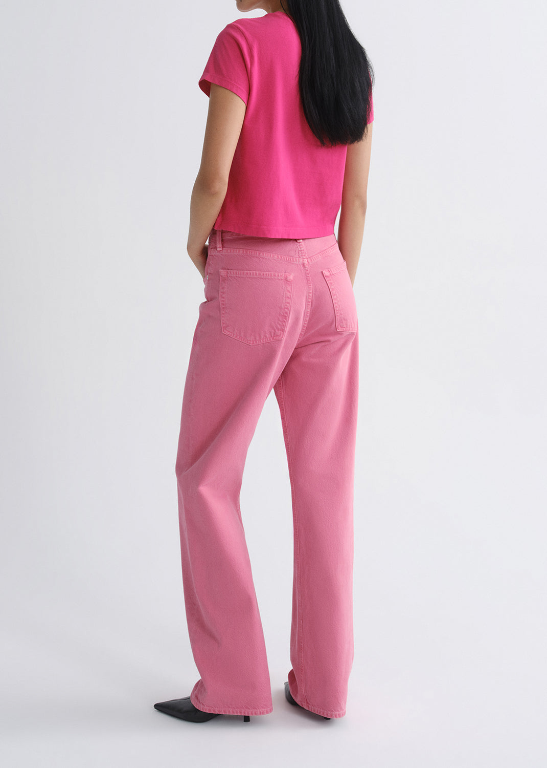 Person wearing a bright pink short sleeve crewneck tee with light pink straight leg jeans viewed from the back on a grey background