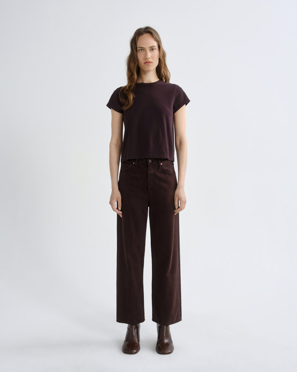A model stands against a light gray studio background wearing a dark plum short-sleeve t-shirt, dark brown wide-leg corduroy pants, and dark brown leather boots.