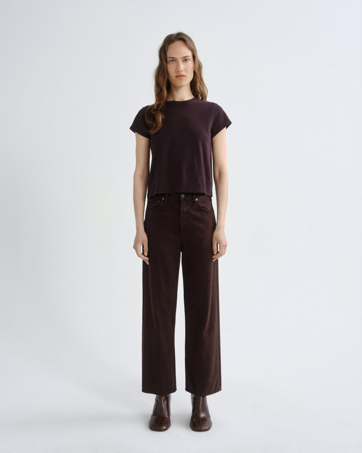 A model stands against a light gray studio background wearing a dark plum short-sleeve t-shirt, dark brown wide-leg corduroy pants, and dark brown leather boots.