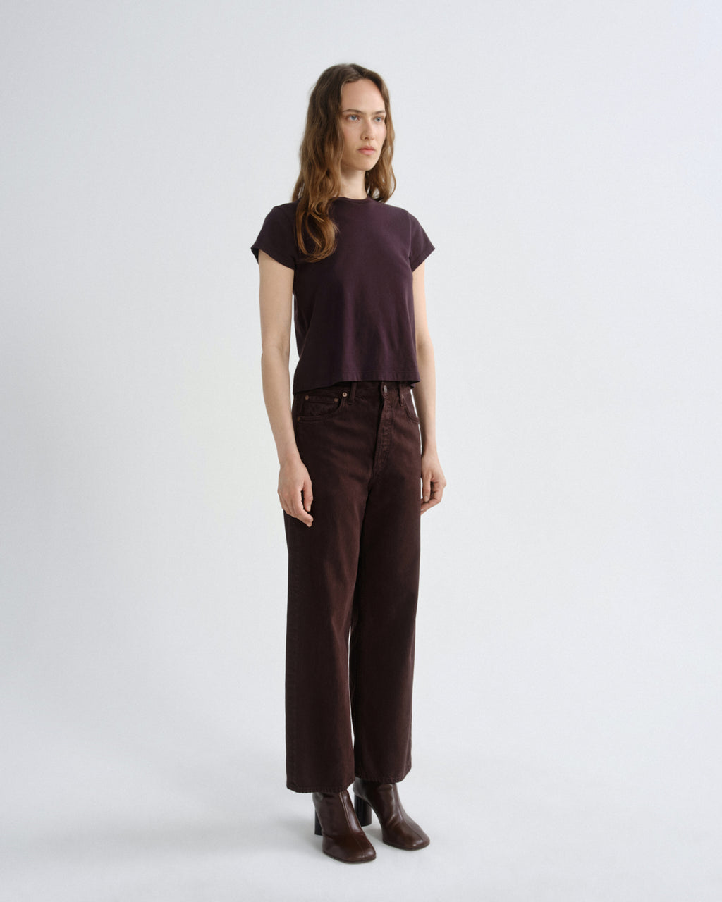 A woman modeling a dark plum short-sleeve top and dark brown wide-leg trousers with brown leather ankle boots, standing against a light gray studio background.