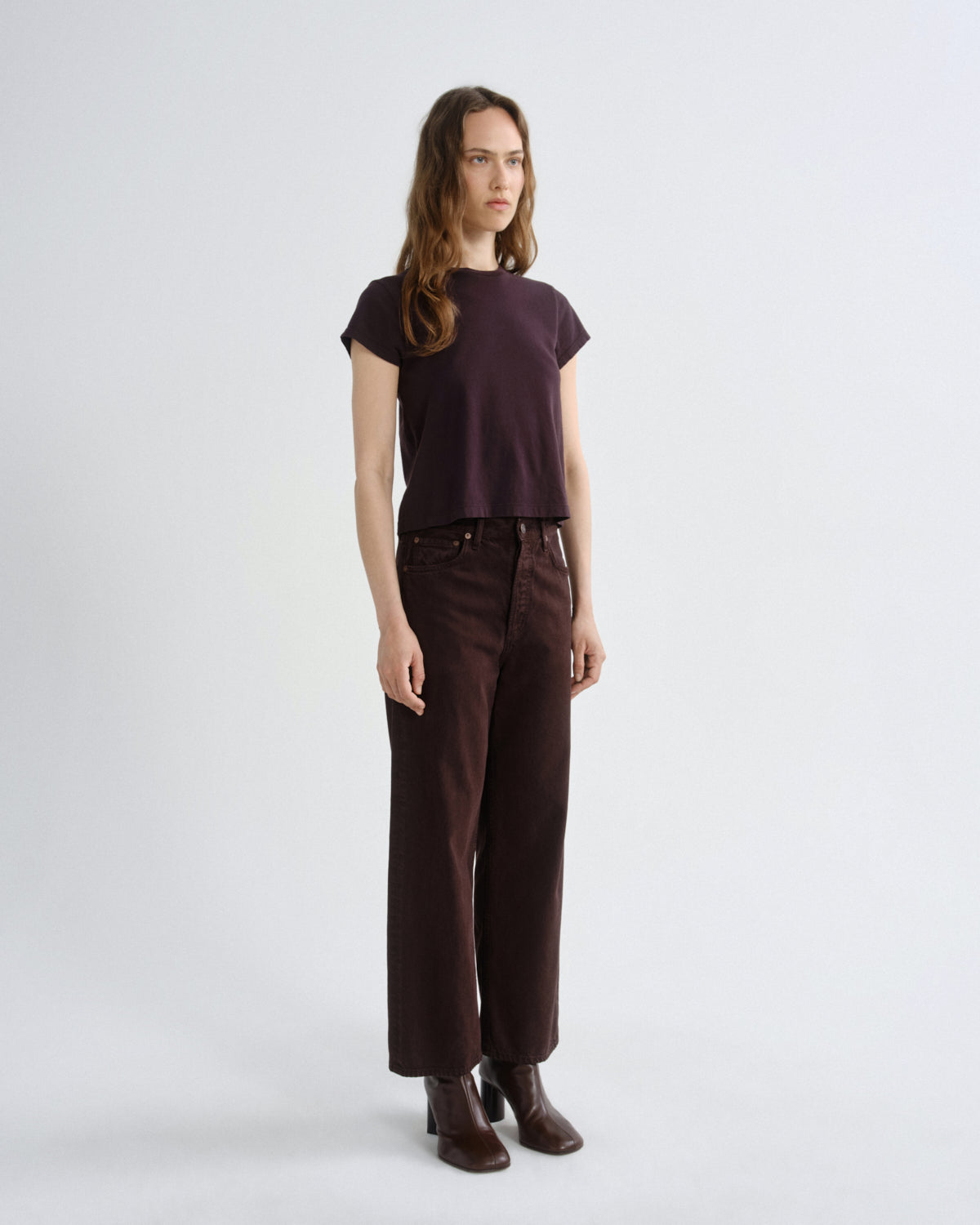 A woman modeling a dark plum short-sleeve top and dark brown wide-leg trousers with brown leather ankle boots, standing against a light gray studio background.