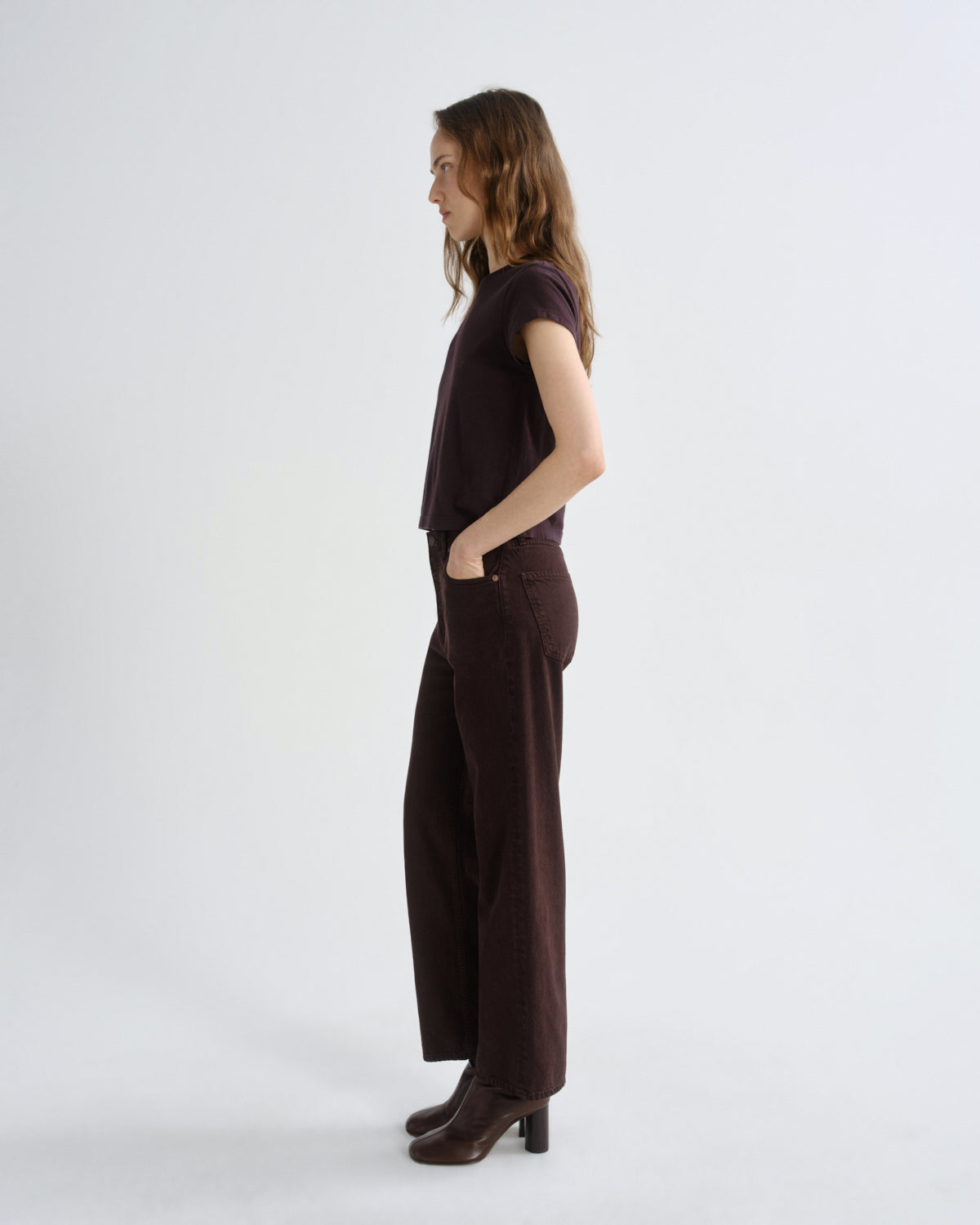 A woman stands in profile wearing a dark brown short-sleeve top and matching wide-leg trousers with dark brown heeled boots, photographed against a clean light gray studio background.