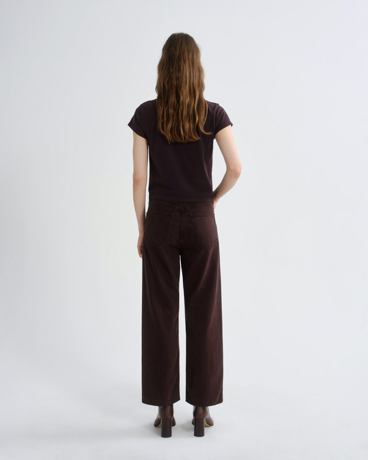 A woman viewed from behind wearing a dark brown short-sleeve t-shirt and matching dark brown wide-leg trousers, standing against a plain light gray studio background.
