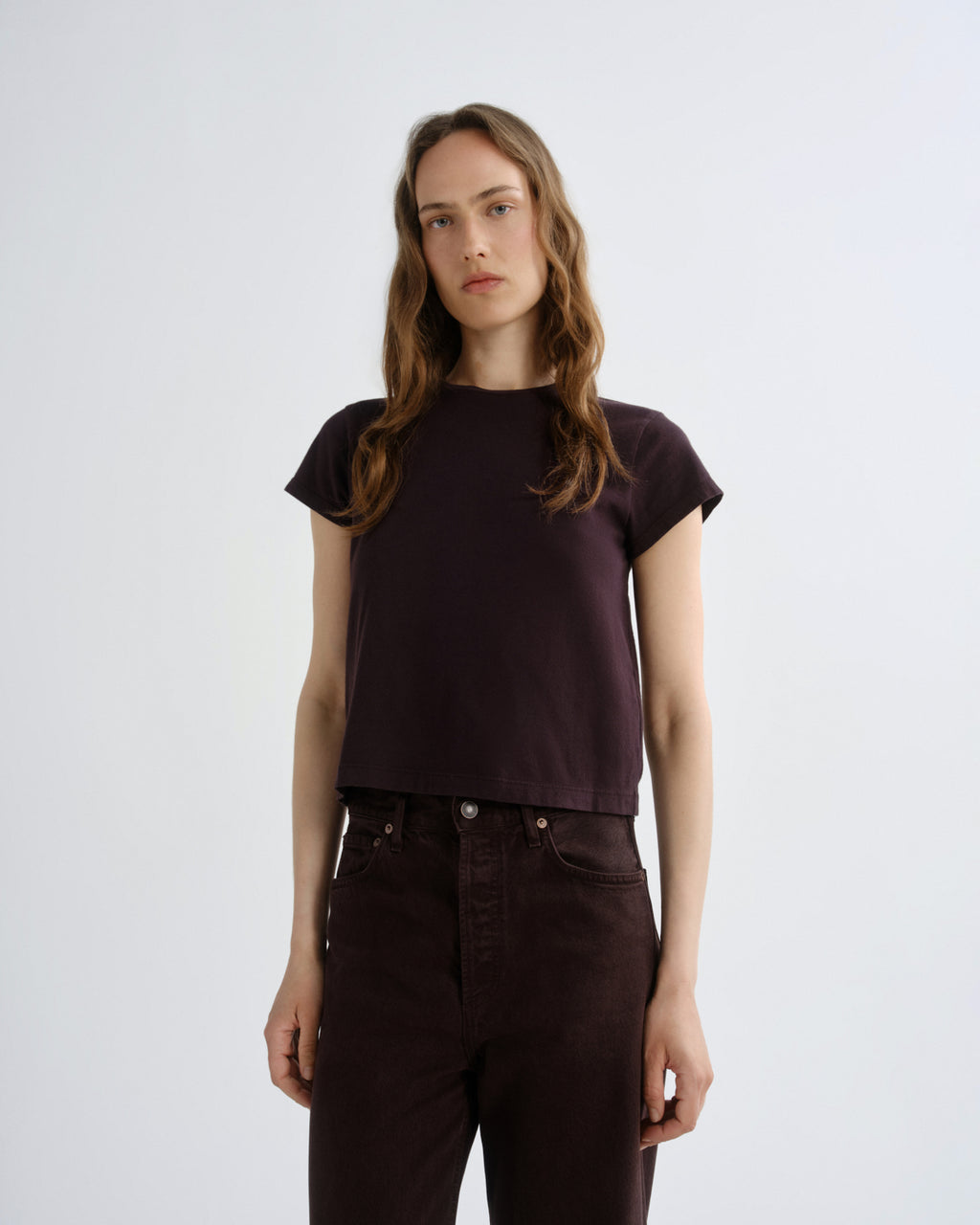 A woman modeling a dark plum short-sleeve crop top with dark brown pants, standing against a light gray studio background.