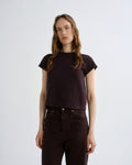 A woman modeling a dark plum short-sleeve crop top with dark brown pants, standing against a light gray studio background.