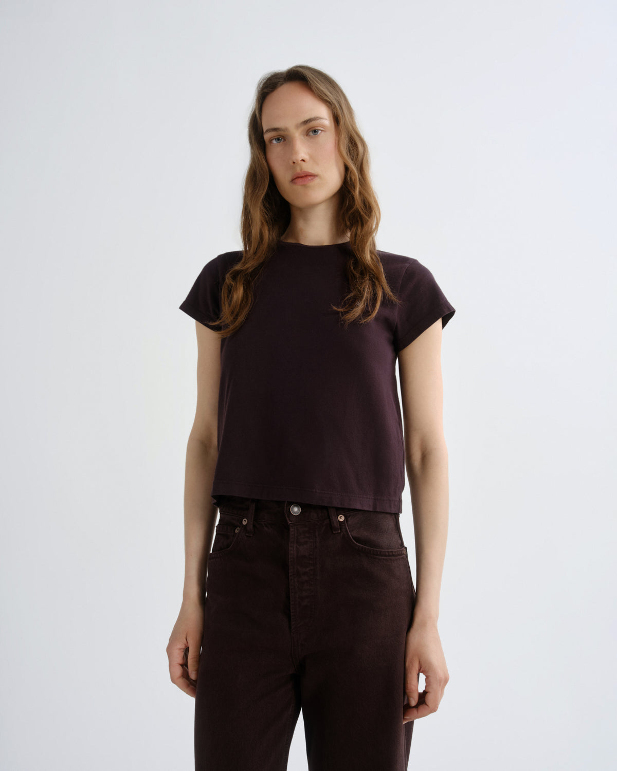A woman modeling a dark plum short-sleeve crop top with dark brown pants, standing against a light gray studio background.