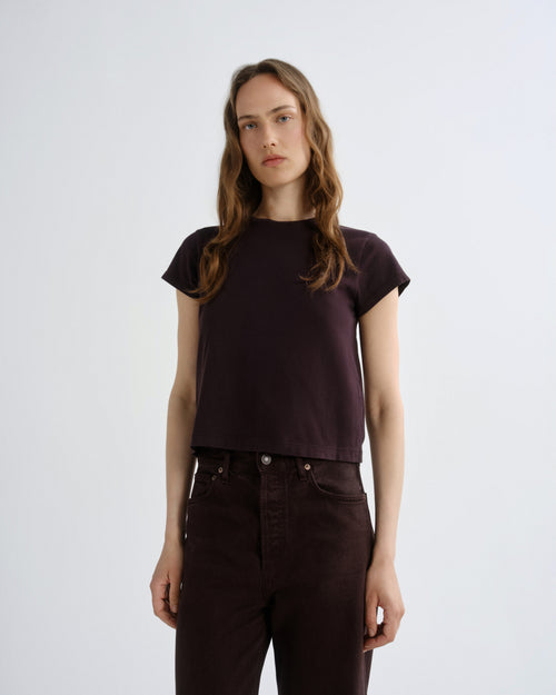 A woman modeling a dark plum short-sleeve crop top with dark brown pants, standing against a light gray studio background.