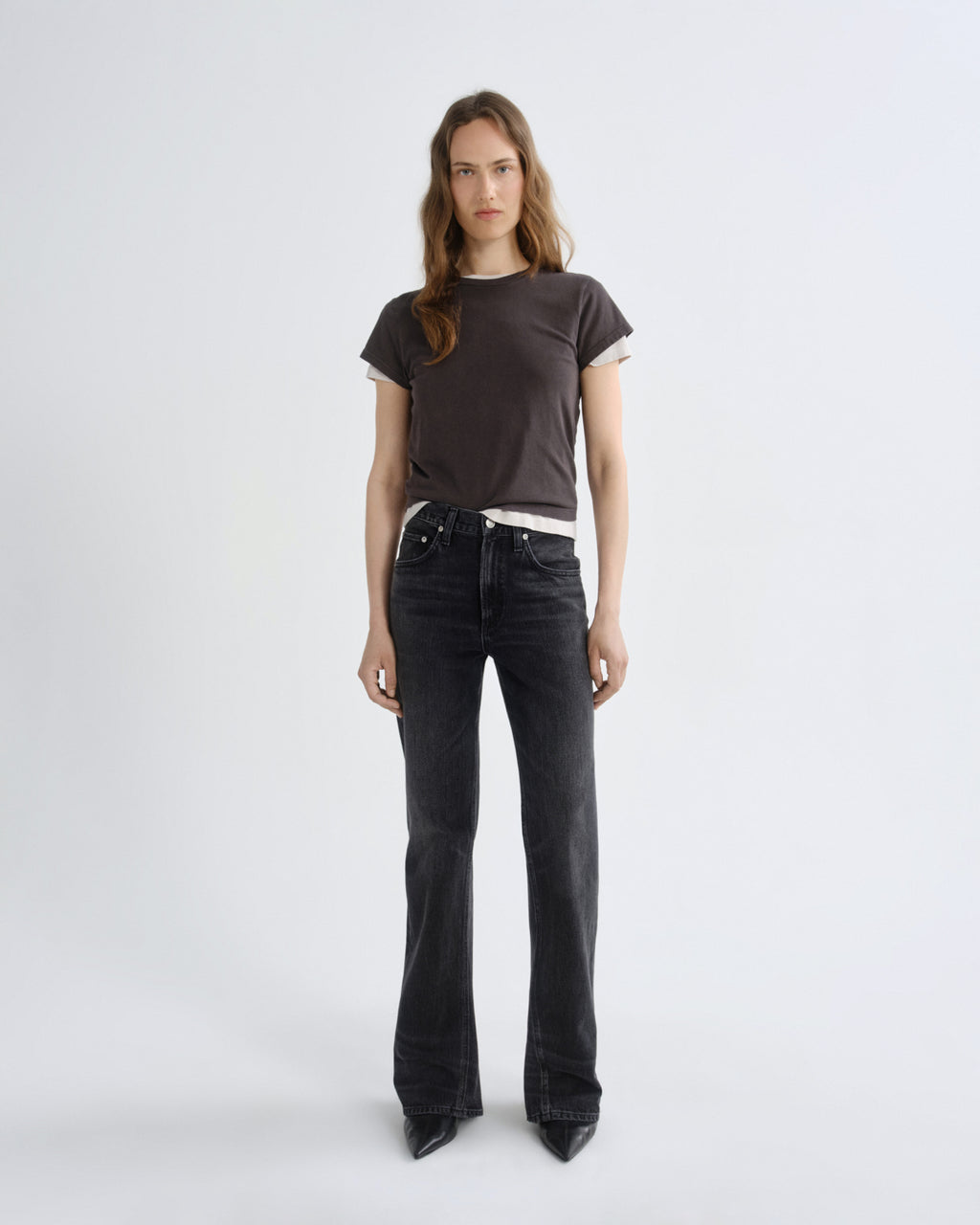 A woman stands against a plain light gray studio background wearing a dark brown short-sleeve t-shirt with white ribbed trim, black wide-leg jeans, and black pointed shoes.