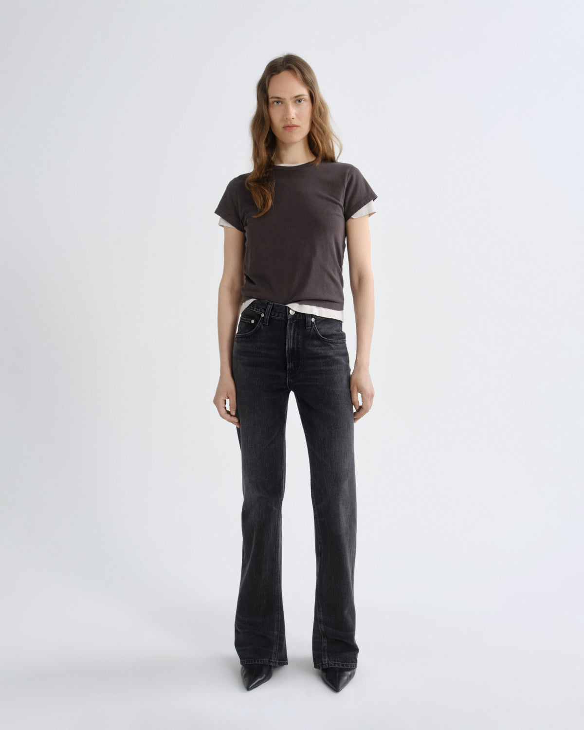 A woman stands against a plain light gray studio background wearing a dark brown short-sleeve t-shirt with white ribbed trim, black wide-leg jeans, and black pointed shoes.