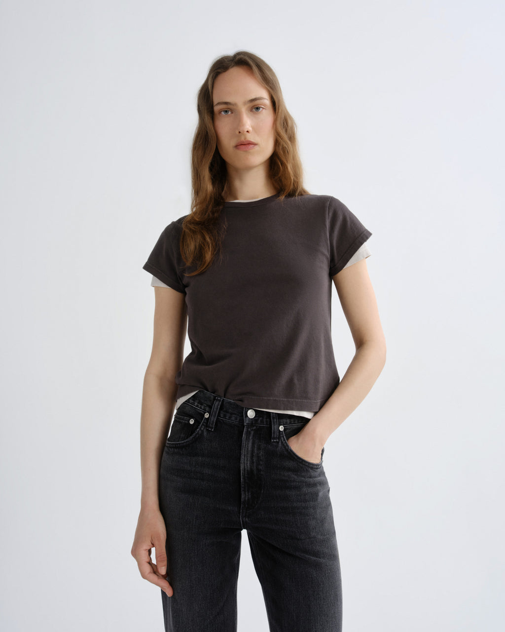 A woman modeling a dark brown short-sleeve t-shirt with a subtle white inner collar, paired with dark gray jeans, against a plain light gray background.