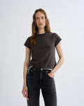 A woman modeling a dark brown short-sleeve t-shirt with a subtle white inner collar, paired with dark gray jeans, against a plain light gray background.