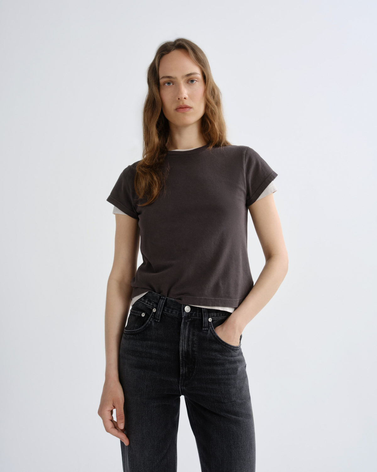 A woman modeling a dark brown short-sleeve t-shirt with a subtle white inner collar, paired with dark gray jeans, against a plain light gray background.
