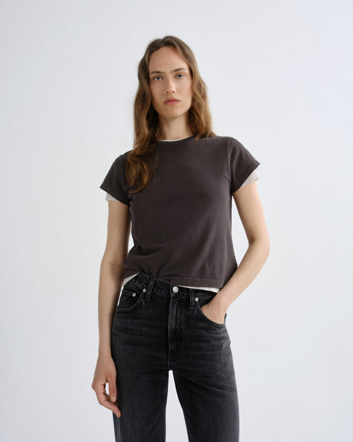 A woman modeling a dark brown short-sleeve t-shirt with a subtle white inner collar, paired with dark gray jeans, against a plain light gray background.