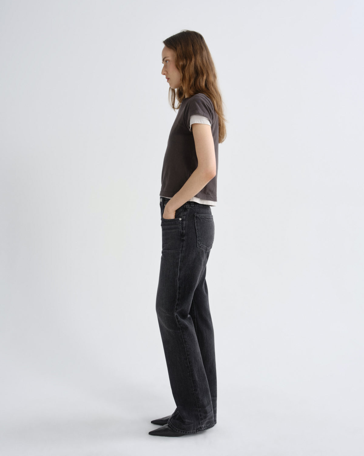 A model standing in profile wearing a dark brown cropped t-shirt with a white undershirt, dark gray wide-leg jeans, and black pointed shoes against a light gray studio background.
