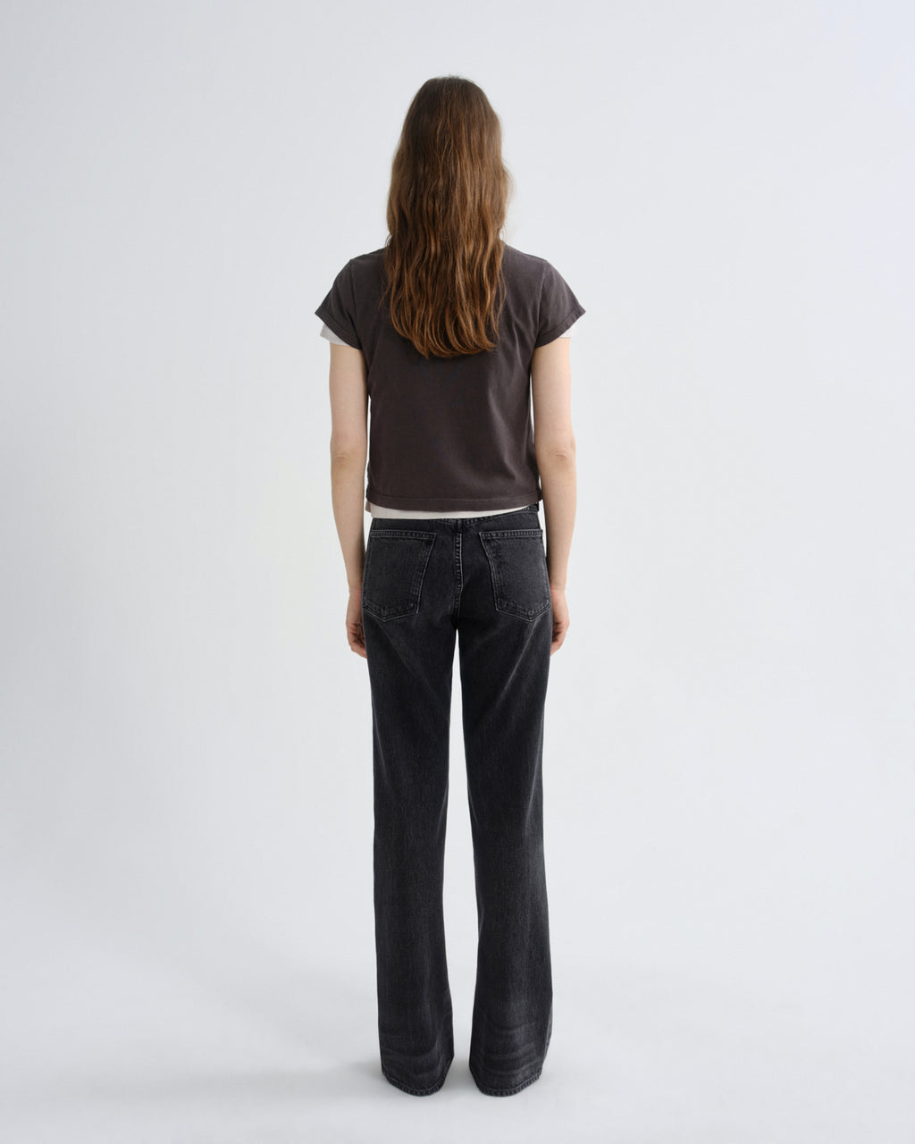 A woman viewed from behind wearing a dark brown short-sleeve t-shirt and dark charcoal gray wide-leg jeans, standing against a light gray studio background.