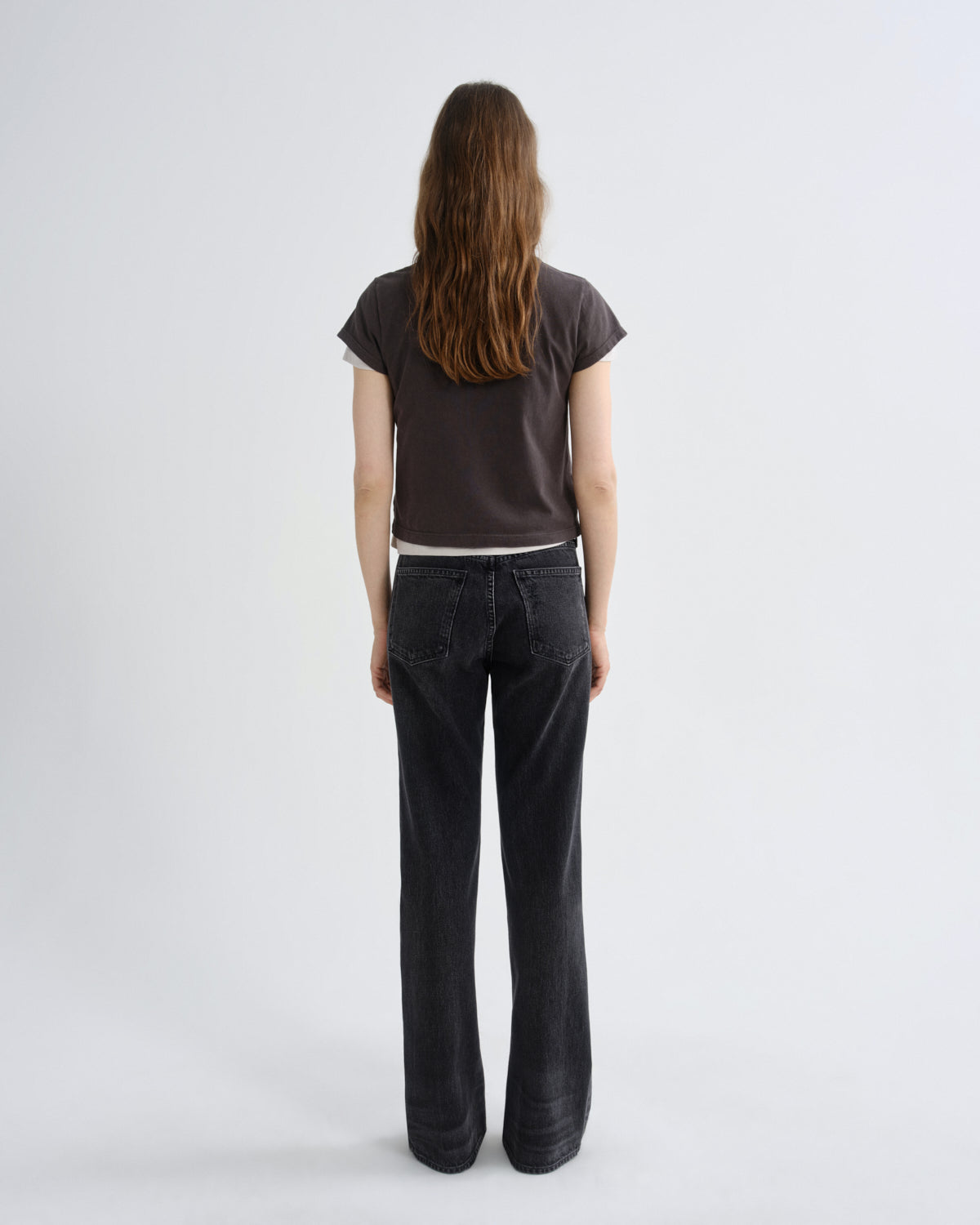 A woman viewed from behind wearing a dark brown short-sleeve t-shirt and dark charcoal gray wide-leg jeans, standing against a light gray studio background.