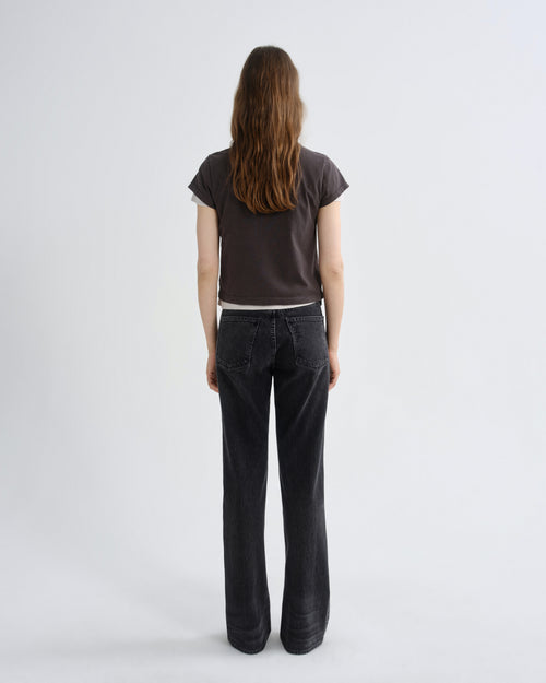 A woman viewed from behind wearing a dark brown short-sleeve t-shirt and dark charcoal gray wide-leg jeans, standing against a light gray studio background.