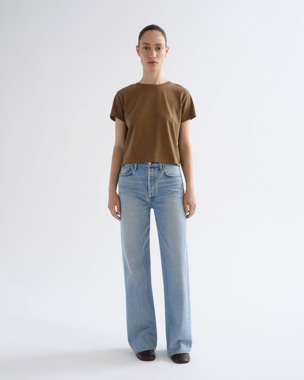 A model stands against a light gray studio background wearing a short-sleeve brown t-shirt and light-wash blue wide-leg jeans.