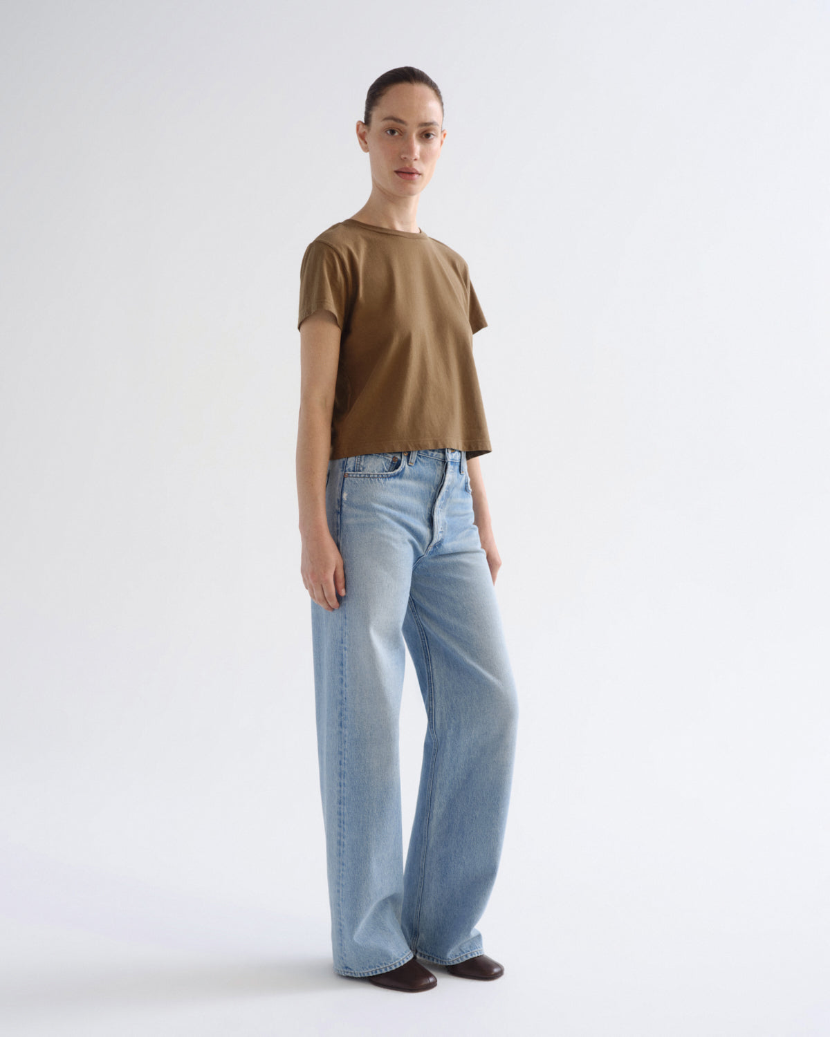 A model stands against a light gray studio background wearing a cropped brown short-sleeve t-shirt and light-wash wide-leg jeans.