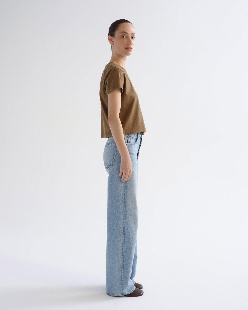 A woman standing in profile wearing a cropped brown short-sleeve top and light blue wide-leg jeans against a plain white studio background.