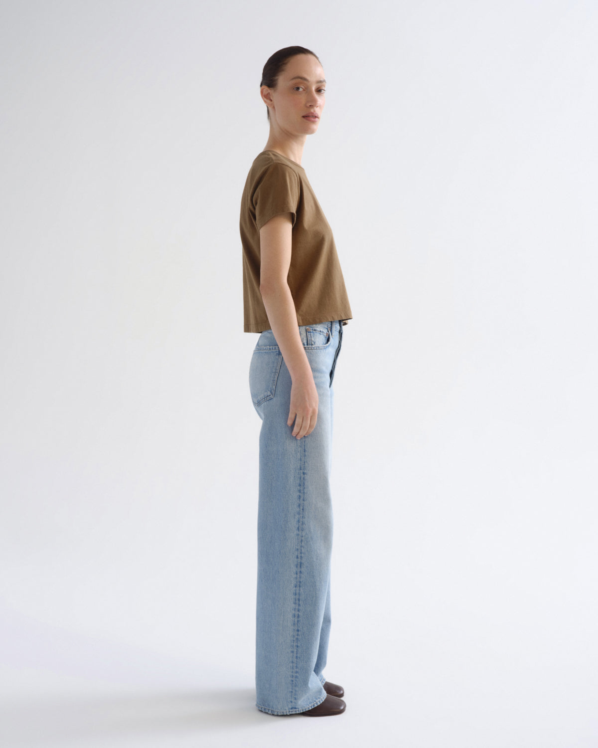 A woman standing in profile wearing a cropped brown short-sleeve top and light blue wide-leg jeans against a plain white studio background.