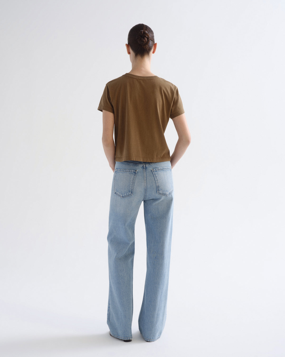 A model viewed from behind wearing a brown short-sleeve t-shirt and light wash wide-leg jeans against a plain white studio background.