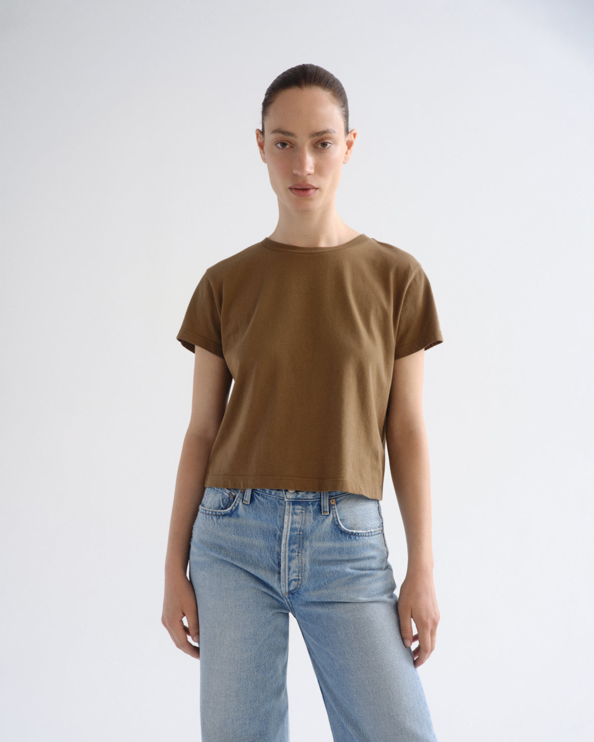 A model wearing a cropped, short-sleeve brown t-shirt and light wash blue jeans, standing against a plain light gray studio background.