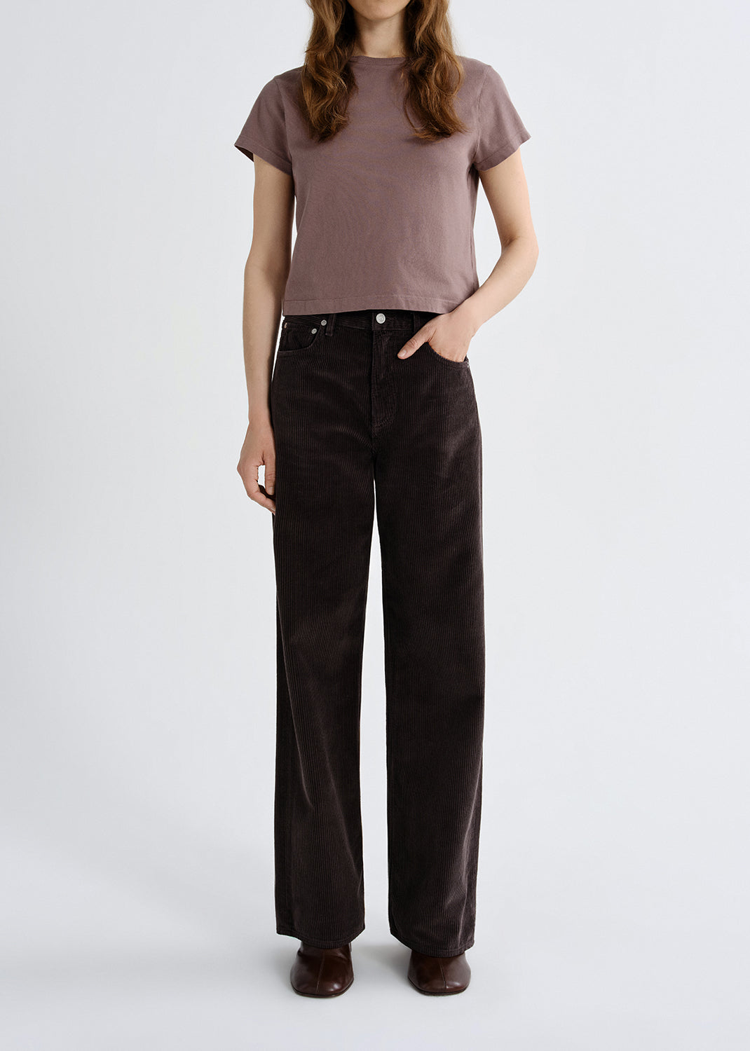 Person wearing a mauve short sleeve crewneck tee with dark brown straight leg jeans on a grey background