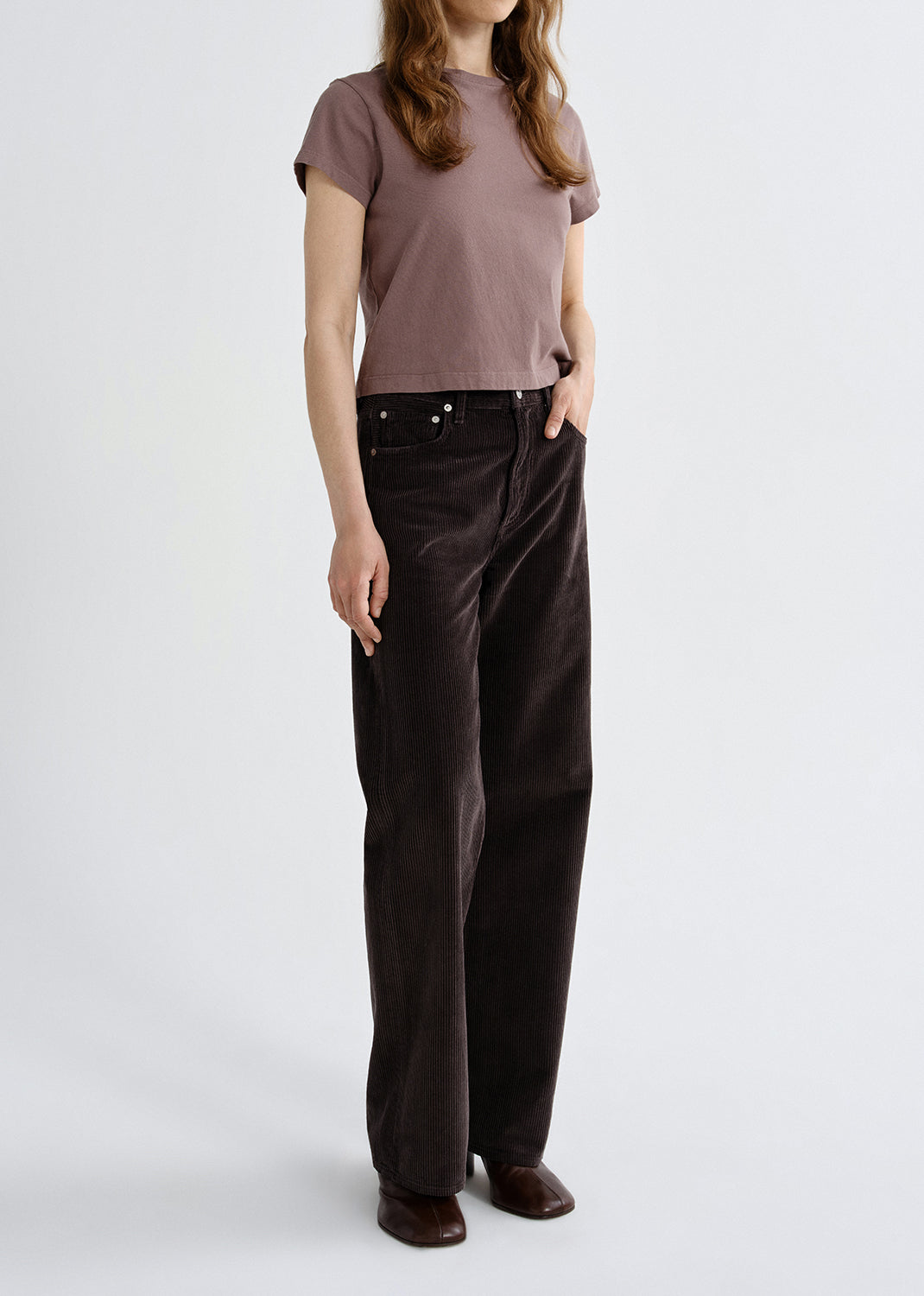Person wearing a mauve short sleeve crewneck tee with dark brown straight leg jeans viewed from the side on a grey background