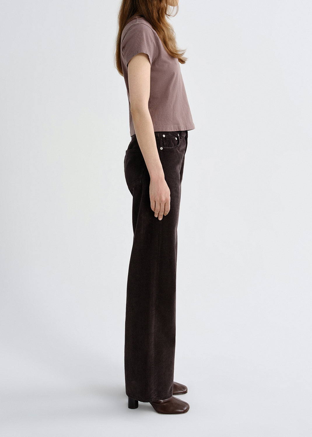Person wearing a mauve short sleeve crewneck tee with dark brown straight leg jeans viewed from the side on a grey background