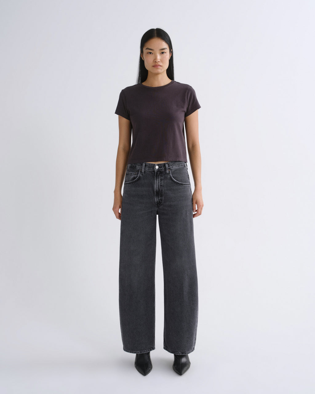 A woman modeling a dark grey short-sleeve crop top paired with dark gray jeans, standing against a light gray studio background.