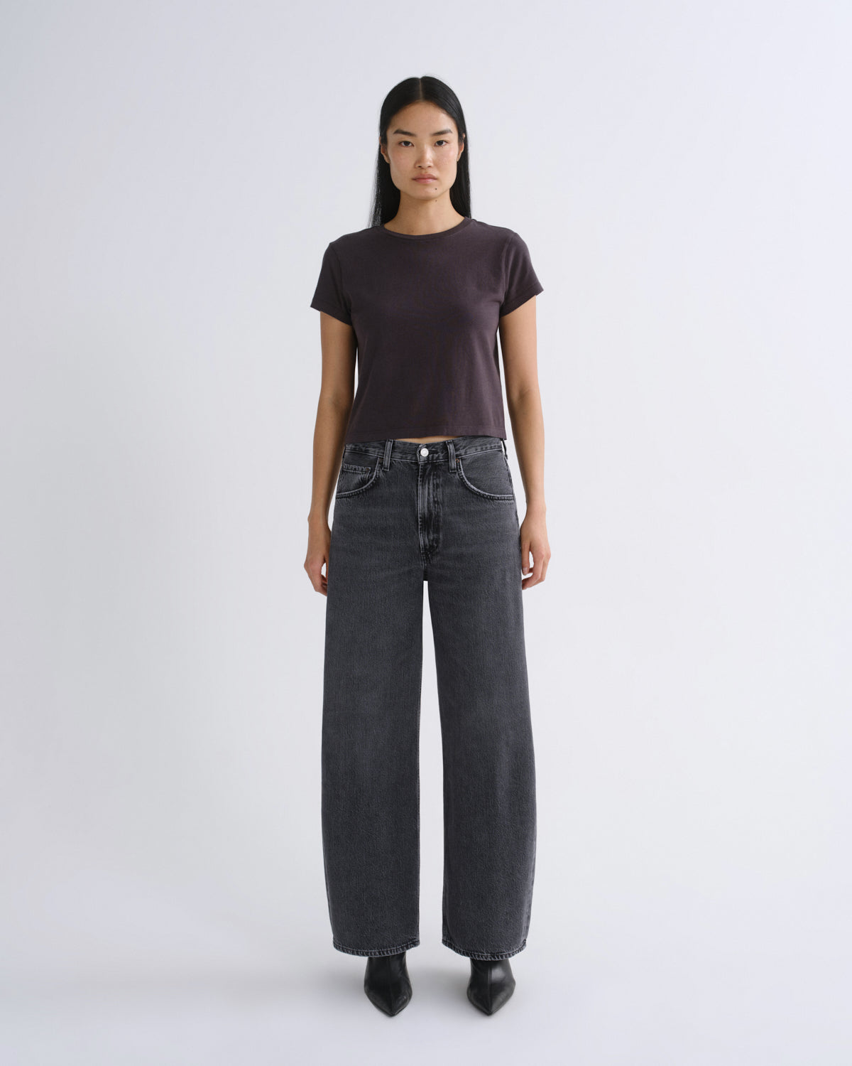 A woman modeling a dark grey short-sleeve crop top paired with dark gray jeans, standing against a light gray studio background.