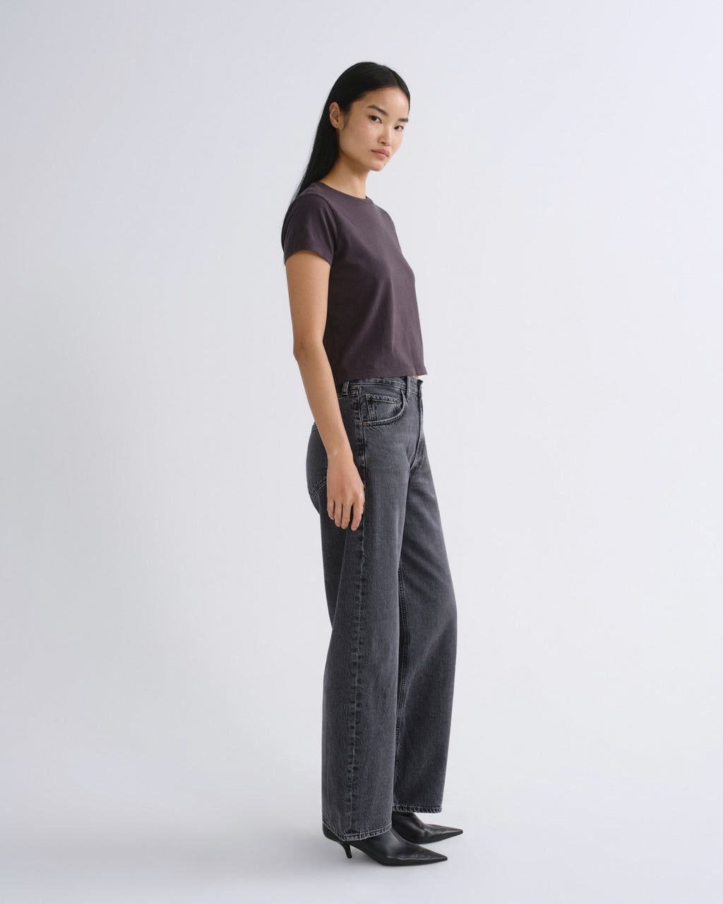A woman modeling a dark grey short-sleeve crop top paired with dark gray jeans, standing against a light gray studio background.