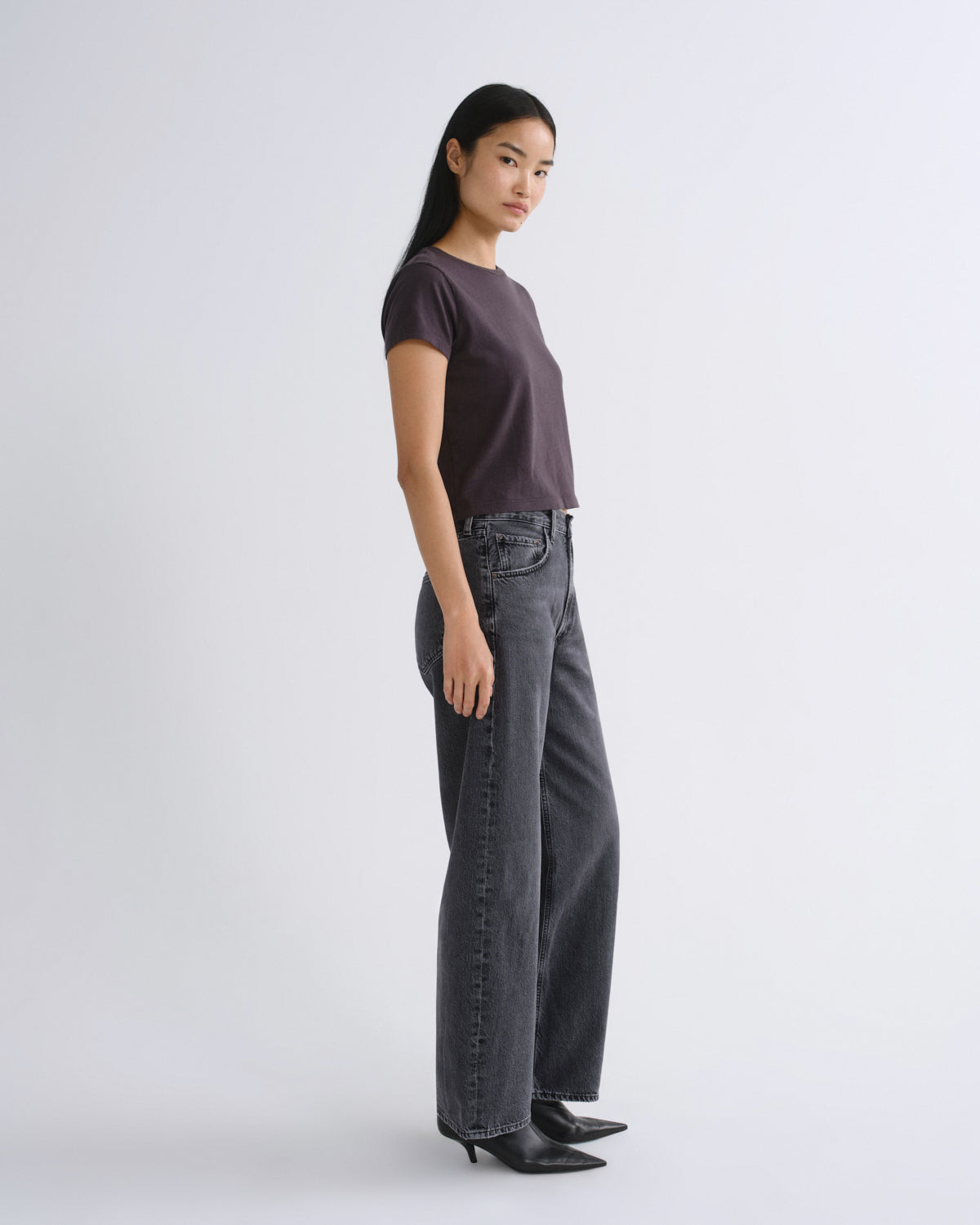 A woman modeling a dark grey short-sleeve crop top paired with dark gray jeans, standing against a light gray studio background.