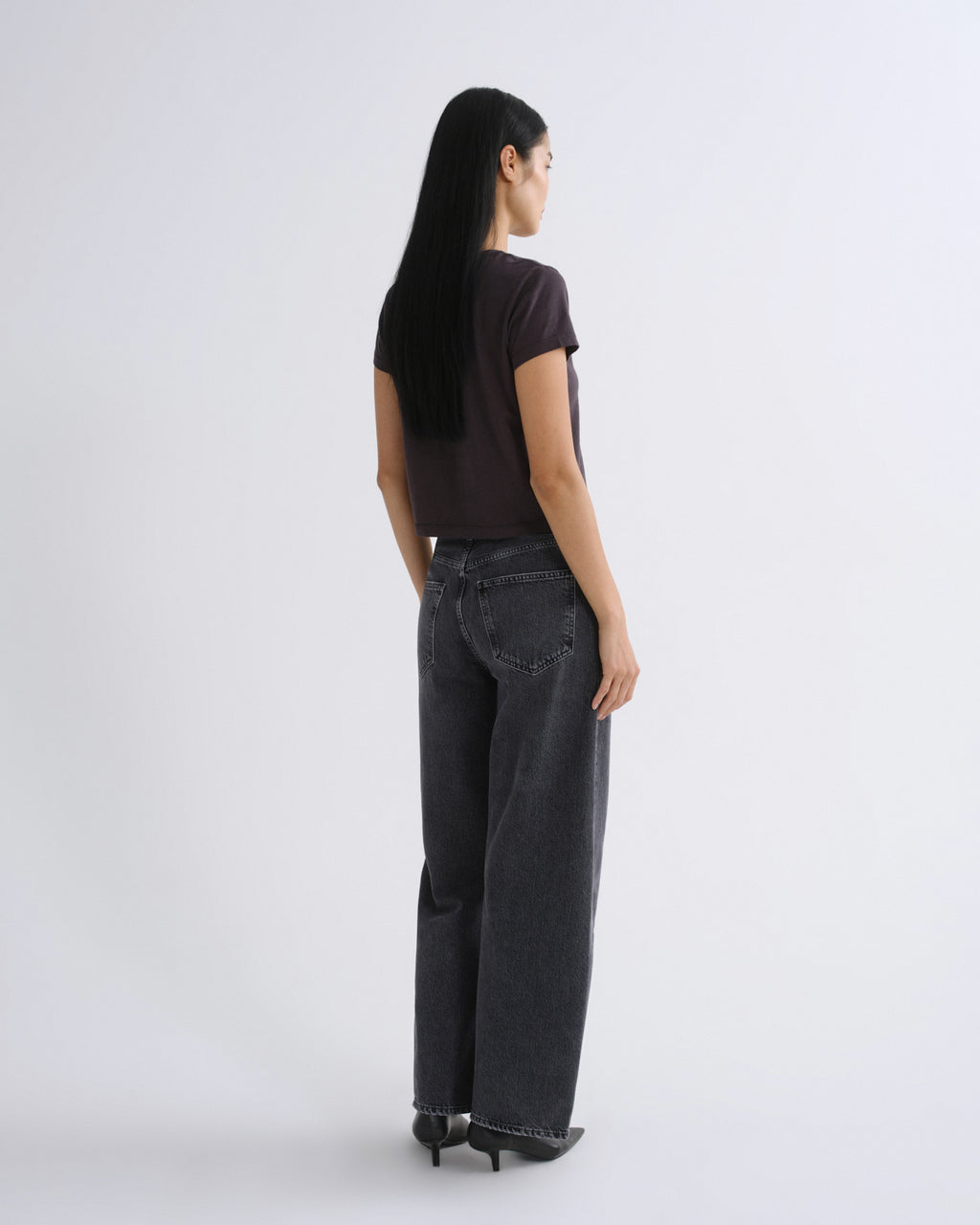A woman modeling a dark grey short-sleeve crop top paired with dark gray jeans, standing against a light gray studio background.