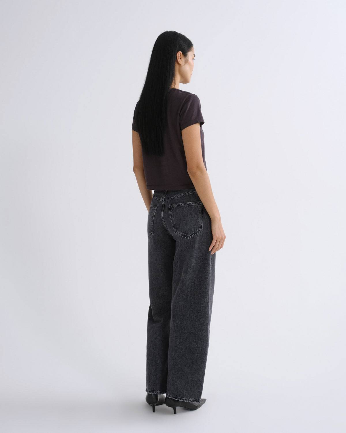 A woman modeling a dark grey short-sleeve crop top paired with dark gray jeans, standing against a light gray studio background.