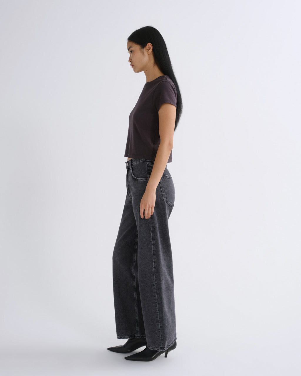 A woman modeling a dark grey short-sleeve crop top paired with dark gray jeans, standing against a light gray studio background.