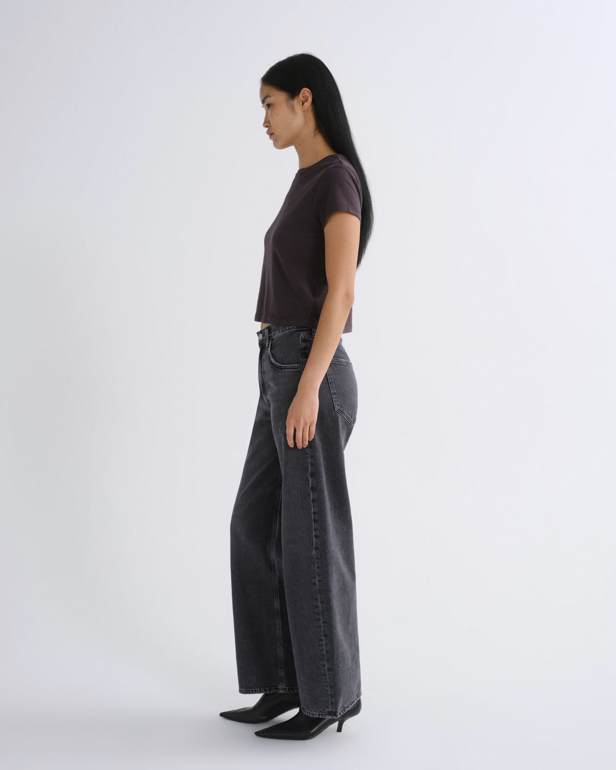 A woman modeling a dark grey short-sleeve crop top paired with dark gray jeans, standing against a light gray studio background.