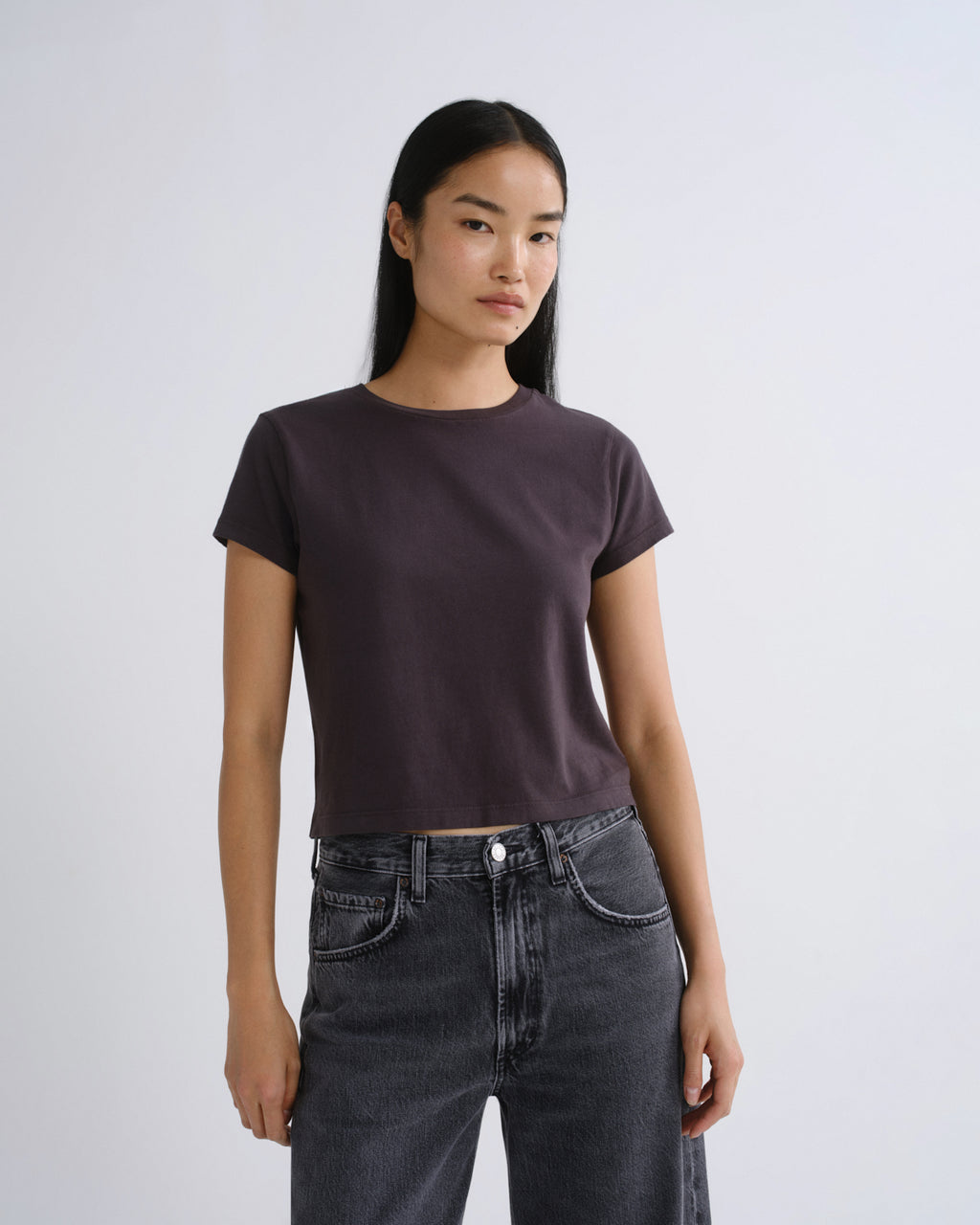 A woman modeling a dark grey short-sleeve crop top paired with dark gray jeans, standing against a light gray studio background.