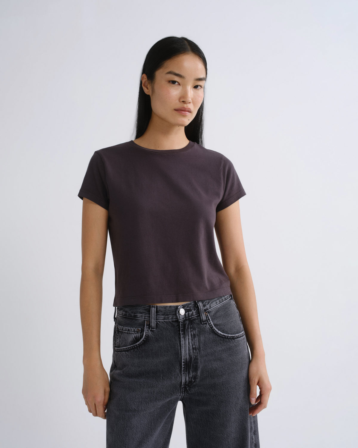 A woman modeling a dark grey short-sleeve crop top paired with dark gray jeans, standing against a light gray studio background.