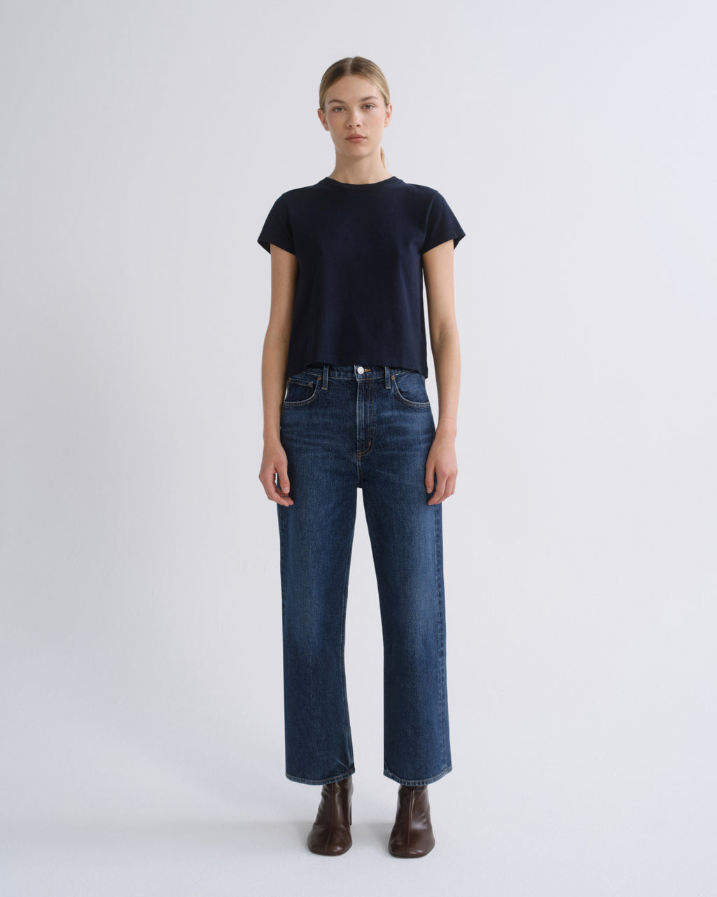 A model stands against a light gray studio background wearing a dark navy short-sleeve crewneck t-shirt, dark blue straight-leg jeans, and dark brown leather ankle boots.