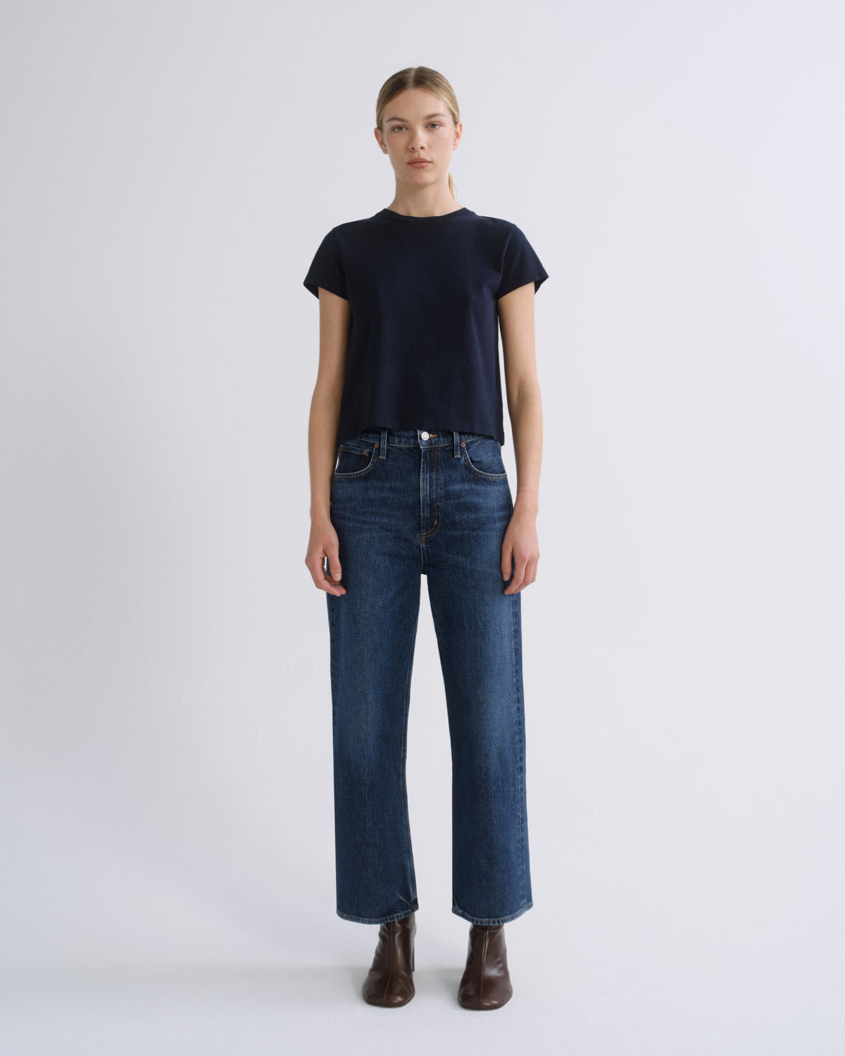 A model stands against a light gray studio background wearing a dark navy short-sleeve crewneck t-shirt, dark blue straight-leg jeans, and dark brown leather ankle boots.