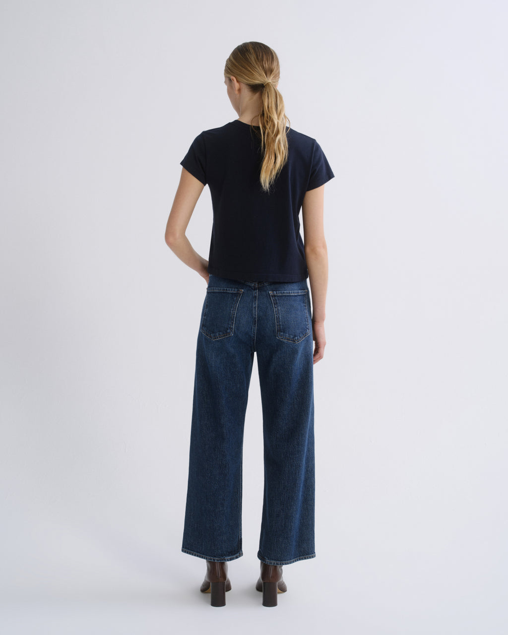 A woman wearing a dark navy short-sleeve top, blue straight-leg jeans, and brown heeled ankle boots, standing against a light gray studio background.