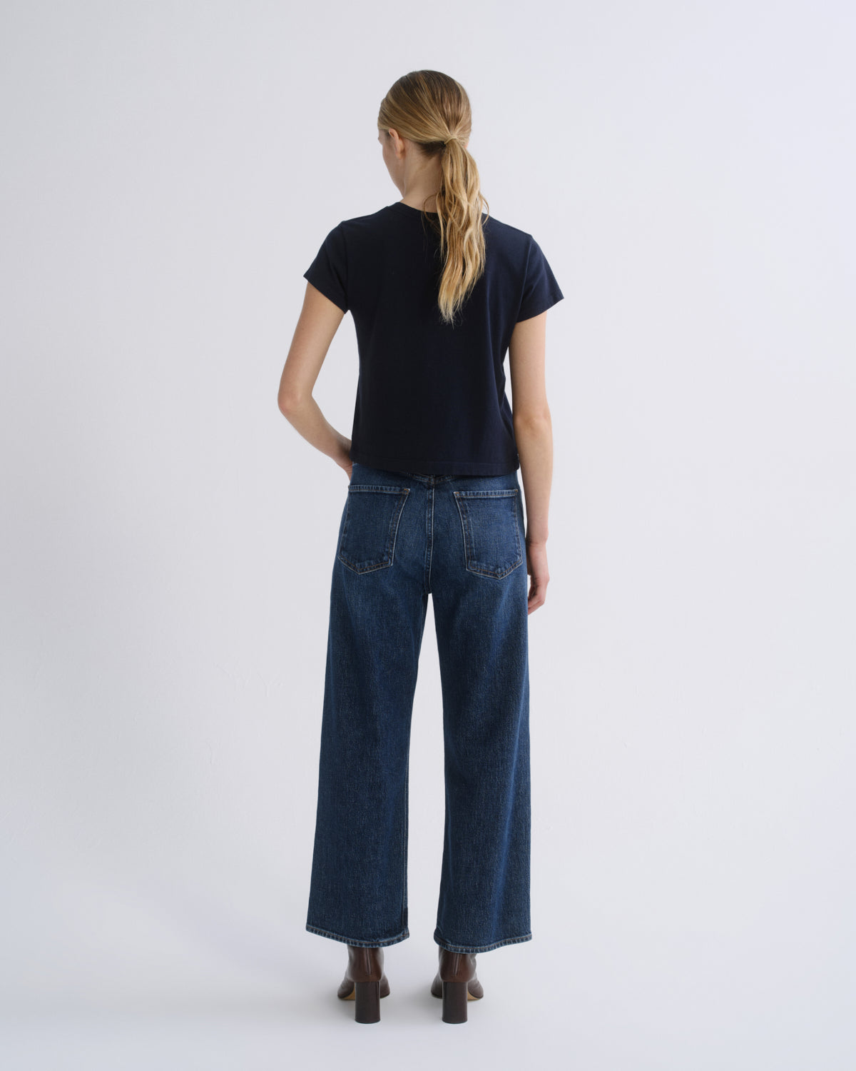 A woman wearing a dark navy short-sleeve top, blue straight-leg jeans, and brown heeled ankle boots, standing against a light gray studio background.