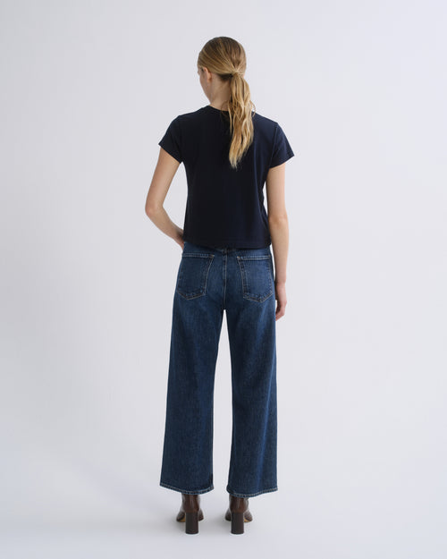 A woman wearing a dark navy short-sleeve top, blue straight-leg jeans, and brown heeled ankle boots, standing against a light gray studio background.