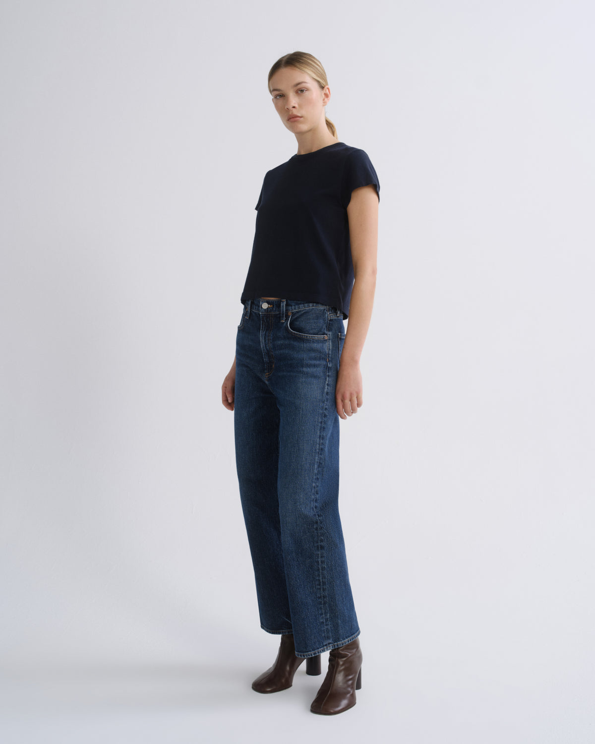 A woman modeling a dark navy short-sleeve knit top and dark blue wide-leg jeans with brown ankle boots, standing against a light gray studio background.