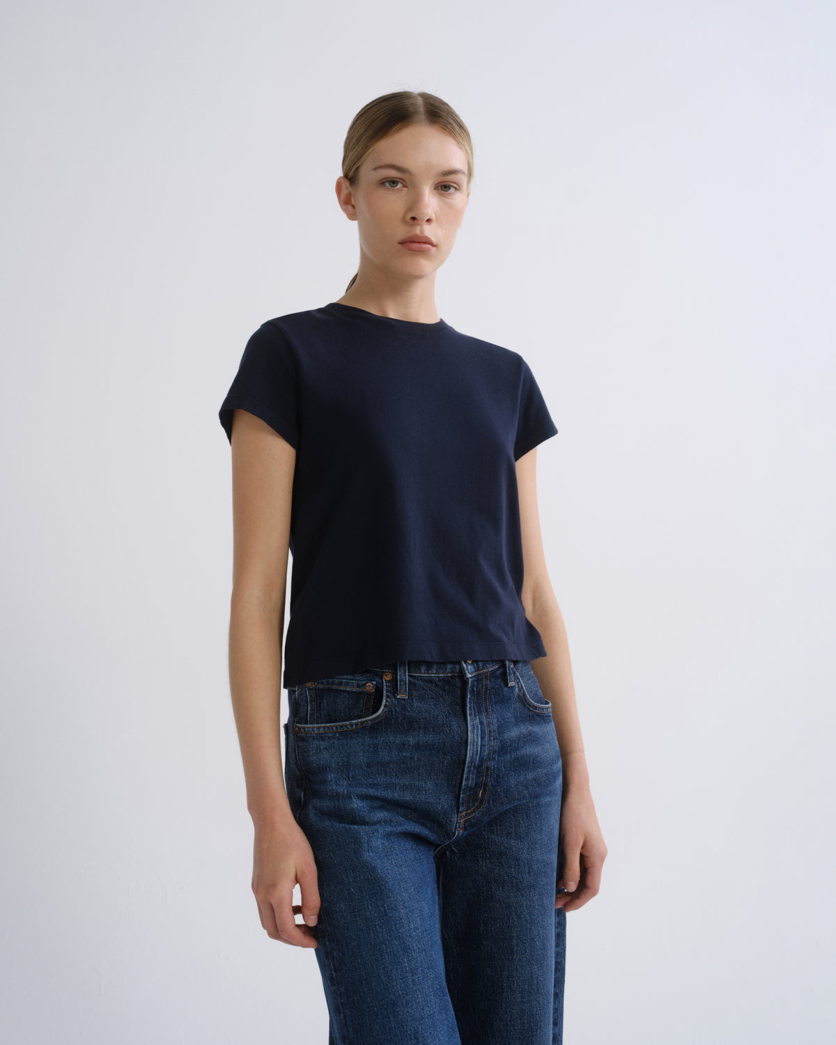 A woman modeling a dark navy blue short-sleeve crewneck t-shirt paired with blue jeans, standing against a light gray studio background.