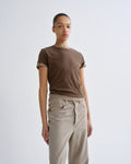 A model wearing a brown short-sleeve crewneck t-shirt with rolled cuffs and beige wide-leg jeans, standing against a light gray studio background.