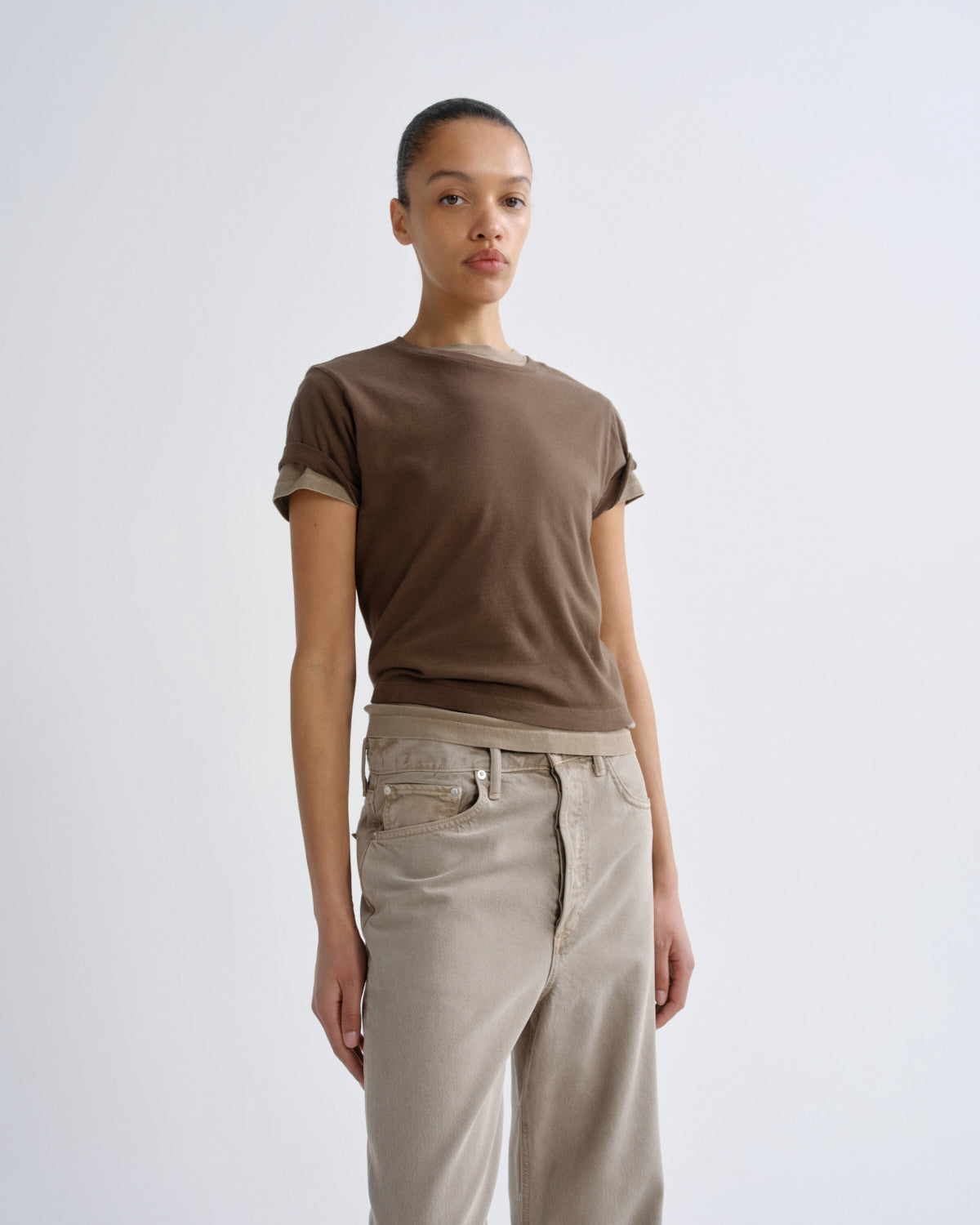 A model wearing a brown short-sleeve crewneck t-shirt with rolled cuffs and beige wide-leg jeans, standing against a light gray studio background.