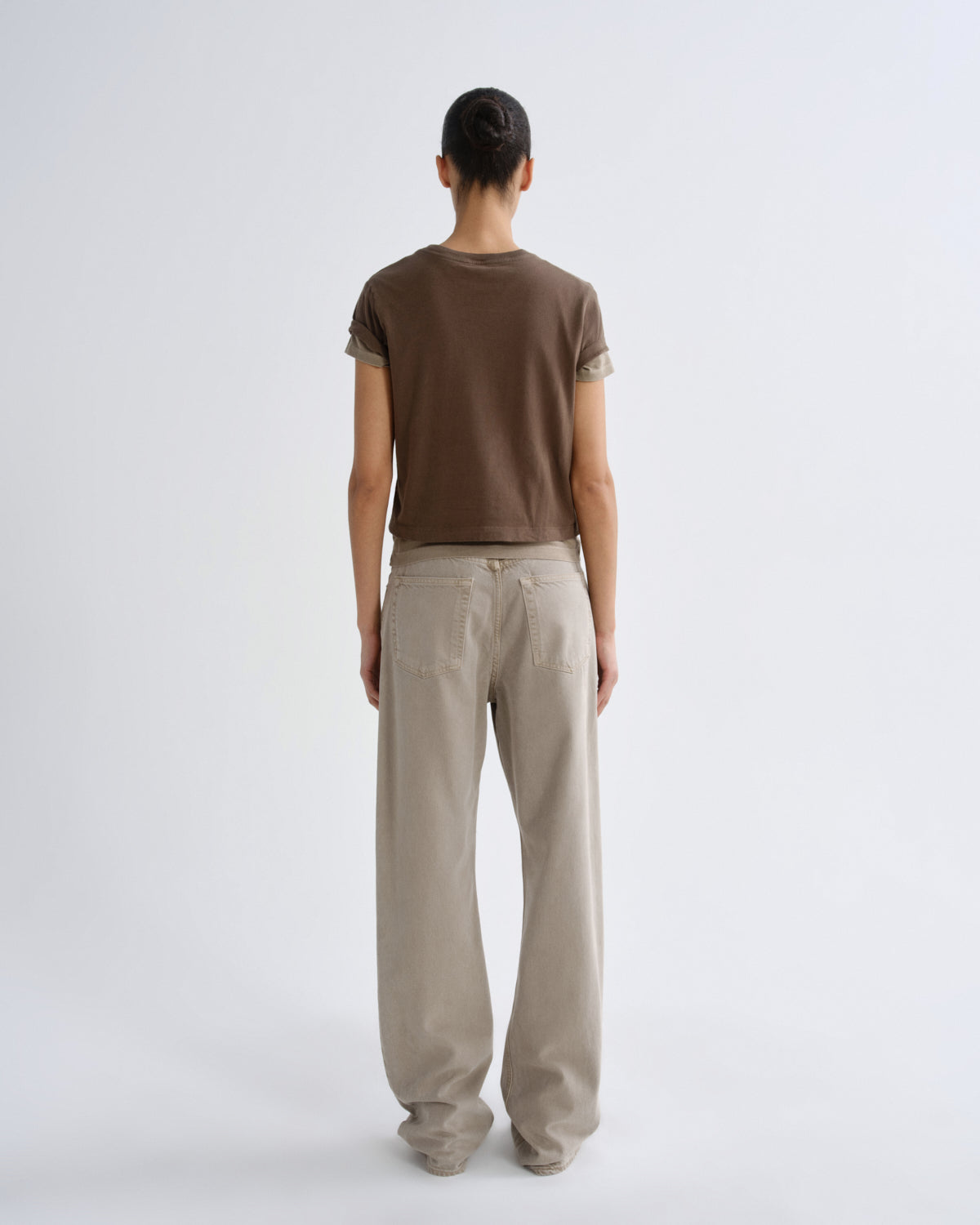A model viewed from the back wearing a brown short-sleeve t-shirt with rolled-up sleeves and beige wide-leg trousers, set against a plain light gray studio background.